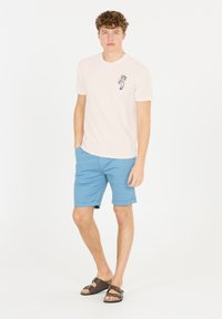Ljust rosa t-shirt med palmträdsmotiv, kombinerad med blå shorts och bruna sandaler. Modellen står mot en vit bakgrund.