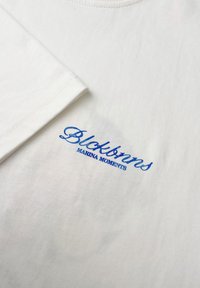 Witte katoenen t-shirt met een geribbeld textuur. Voorzien van blauw geborduurde tekst: "Blockawns" en "MARINA MOMENTS" op de voorkant.