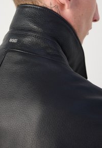 Jachetă din piele neagră cu guler înalt, suprafață texturată și cusături subtile, având un logo argintiu "BOSS" pe guler.