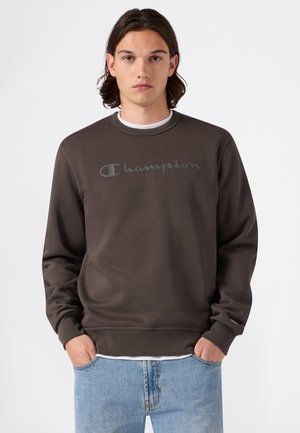 Jeune homme aux cheveux bruns mi-longs porte un sweat-shirt Champion marron foncé et un jean bleu clair, les mains dans les poches, sur fond blanc.