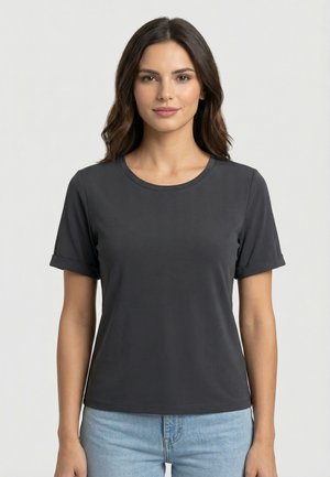 Donna con capelli castani di media lunghezza che indossa una maglietta grigia scura a maniche corte senza decorazioni e jeans azzurri, in piedi davanti a uno sfondo bianco.