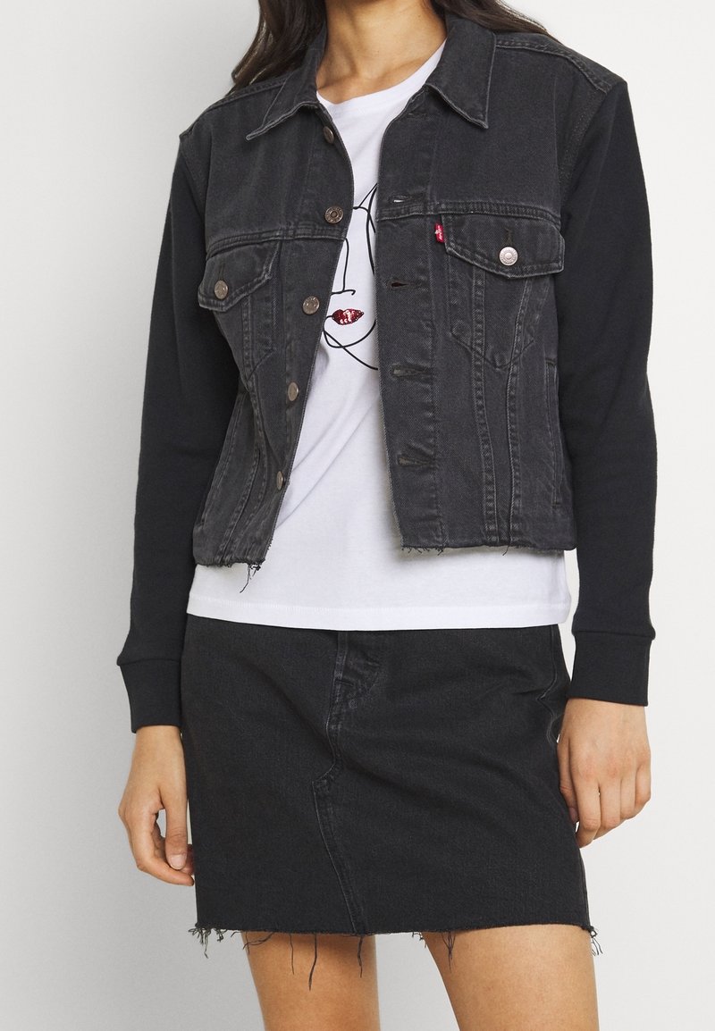 Femme portant une veste en jean noire à manches longues avec boutons, un t-shirt blanc à motif graphique, et une jupe en jean noire avec un ourlet effiloché.