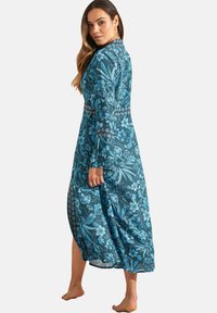 Lang, langmouwig jurk in donker teal met ingewikkelde blauwe bloemenpatronen, voorzien van een kraag en een zijsplit. Zacht, vloeiend materiaal.