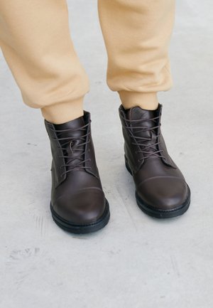 Braune Leder-Stiefel knöchelhochel mit Schnürsenkeln, runder Spitze und strukturiertem Gummisohle, kombiniert mit beigen, umkrempelten Sweatpants.