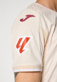 Bežinė sportinė marškinė su raudona "J" logotipu šalia peties ir raudonos bei baltos stilizuotos „47“ ženklo viršuje prie rankogalių ant žmogaus rankos.