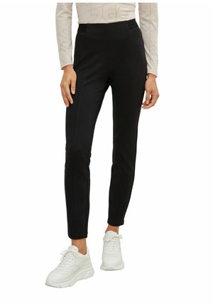 Pantalons noirs ajustés en tissu lisse. Comprend deux poches latérales et un accent à la taille. Associés à des baskets blanches.
