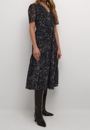 Kvinde iført en midi-kjole med abstrakt mønster i sort og beige med korte ærmer, kombineret med mørkebrune, knæhøje spidse støvler.