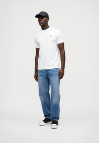 T-shirt branca com um pequeno gráfico no peito, combinado com jeans azul claro e ténis pretos. Boné preto usado ligeiramente torto.