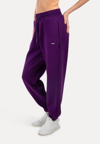 Lila Jogginghose aus weichem Stoff, mit einem engen Bund, Seitentaschen und elastischen Bündchen an den Knöcheln, kombiniert mit weißen Turnschuhen.