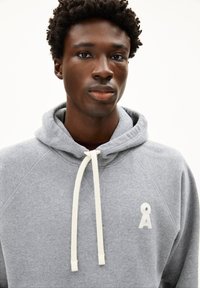 Junger Mann mit kurzen lockigen Haaren, der ein graues Kapuzensweatshirt mit weißen Kordeln und einem weißen Logo auf der Brust trägt.