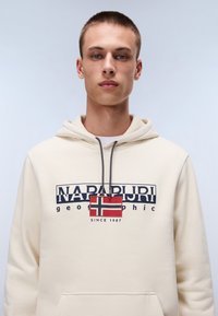 Sudadera crema con diseño frontal, que presenta "NAPAPUR" y una bandera noruega. Incluye un bolsillo tipo canguro y capucha con cordón. Textura suave.
