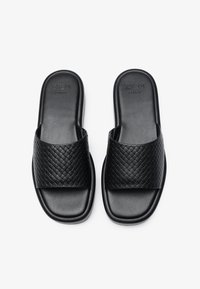 Black slip-on sandals with a woven textured strap, flat sole, and smooth leather finish. Brand logo embossed on the footbed.