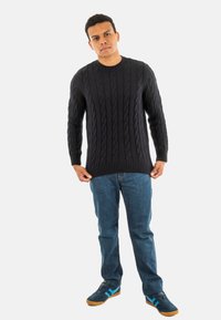 Man staat tegen een witte achtergrond, gekleed in een zwart kabelgebreid sweater, blauwe jeans en marineblauwe sneakers met lichtblauwe strepen.