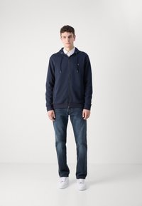Marineblauwe zip-up hoodie met koordjes, gecombineerd met donkerblauwe jeans en witte sneakers. De hoodie heeft een voorkant met rits en een logo op de borst.