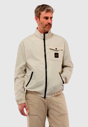 Uomo con capelli grigi che indossa una giacca beige chiusa con cerniera nera e dettagli sulle tasche, abbinata a pantaloni cargo beige.