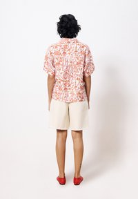 Korte mouw shirt met een roze en oranje botanisch patroon, gecombineerd met beige shorts en rode platte schoenen, gezien van achter.