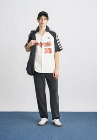 Jovem a usar uma t-shirt preta e branca "Originals 33", calças pretas, ténis brancos e boné preto, segurando uma mala de ombro preta.