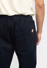 Close-up van een persoon die zwarte broek met elastische tailleband draagt, met een achterzak voorzien van een patch en een klein rood label aan de zaknaad.