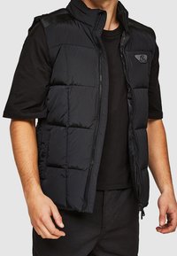 Schwarze gesteppte Weste mit hohem Kragen, Reißverschluss und Seitentaschen. Verfügt über eine gesteppte Textur und ein kleines Logo auf der oberen linken Brust.