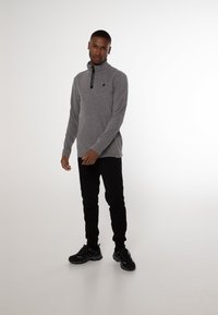 Grå langærmet pullover med højt krave og en sort lynlåsdetalje, parret med sorte tapered bukser og sorte sneakers.