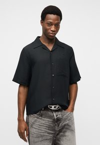 Άνδρας που φοράει κοντομάνικο μαύρο πουκάμισο με κουμπιά και τσέπη στο στήθος, καθώς και ανοιχτό γκρι ξεβαμμένο τζιν με ζώνη ασημένιας αγκράφας.