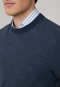 Mornarsko plavi pleteni pulover s okruglim izrezom, složen preko svijetloplavog i bijelog prugastog košulje s ovratnikom. Mekana tekstura i prilagođeni dizajn.