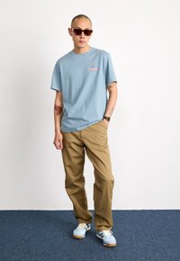 Ljusblå bomulls t-shirt med en liten logotyp, kombinerad med beige byxor och flerfärgade sneakers, kompletterad med bruna solglasögon.