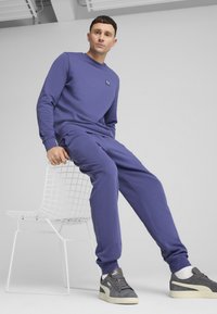 Purple sweatshirt with logo, matching purple sweatpants, and grey sneakers with white accents. Model seated on a white wire chair.