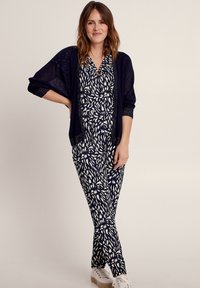 Marine jumpsuit met een wit bladmuster, korte mouwen en een v-hals. Gecombineerd met een bijpassende marine-opengehaakte cardigan en witte sneakers.