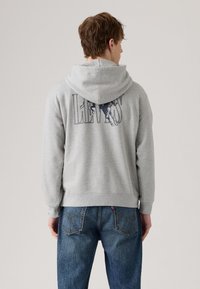 Un sweat à capuche gris avec un grand logo "LEVIS" bleu marine sur le dos, featuring une capuche à cordon et des poignets côtelés, associé à un jean bleu.