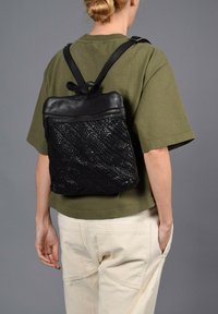 Sac à dos en cuir noir tissé avec un dessus lisse, fermeture zippée et bretelles ajustables, porté sur une chemise verte et un pantalon crème.