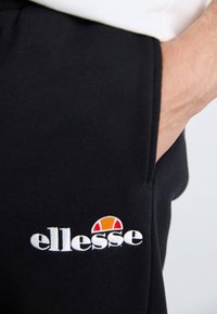 Schwarze Sweatpants mit weichem Material, die ein weißes, gesticktes "ellesse"-Logo und einen farbigen Akzent über dem Logo aufweisen.