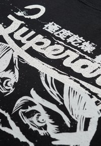 T-shirt nera in cotone con una stampa grafica bianca screpolata di occhi stilizzati e testo. Il design include motivi e texture espressive.