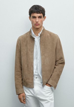 Veste en daim brun clair avec un col haut et une fermeture éclair, associée à une chemise blanche à boutons et un pantalon blanc. Design minimaliste.