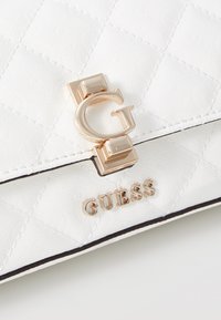Vit kviltad handväska med ett gyllene "G"-låset och guld "GUESS"-logotyp, med en slät textur och svarta accenter längs kanterna.