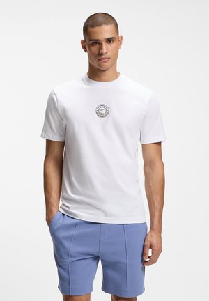 Młody mężczyzna w białej koszulce klubu tenisowego i jasnoniebieskich szortach, stojący z jedną ręką w kieszeni na tle białego tła.