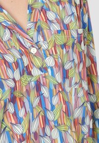 Camisa de colores con cuello, con rayas verticales y patrones de hojas verdes y blancas, que presenta un bolsillo frontal y un solo botón blanco.