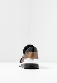 Zapatilla deportiva negra y bronce con un upper texturizado, acentos brillantes, una mediasuela blanca y suela negra. Cuenta con un detalle de marca en la parte trasera.