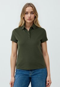 Polo shirt verde scuro con colletto, maniche corte e una patta con bottoni. Abbinato a jeans blu. Tessuto dalla consistenza liscia.