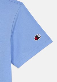 Ljusblå kortärmad t-shirt i bomull, med en broderad logotyp i rött och svart på vänster ärm. Slät textur.