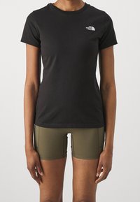 Persona che indossa una t-shirt nera aderente di The North Face e pantaloni corti sportivi verde oliva, in piedi contro uno sfondo chiaro e semplice.