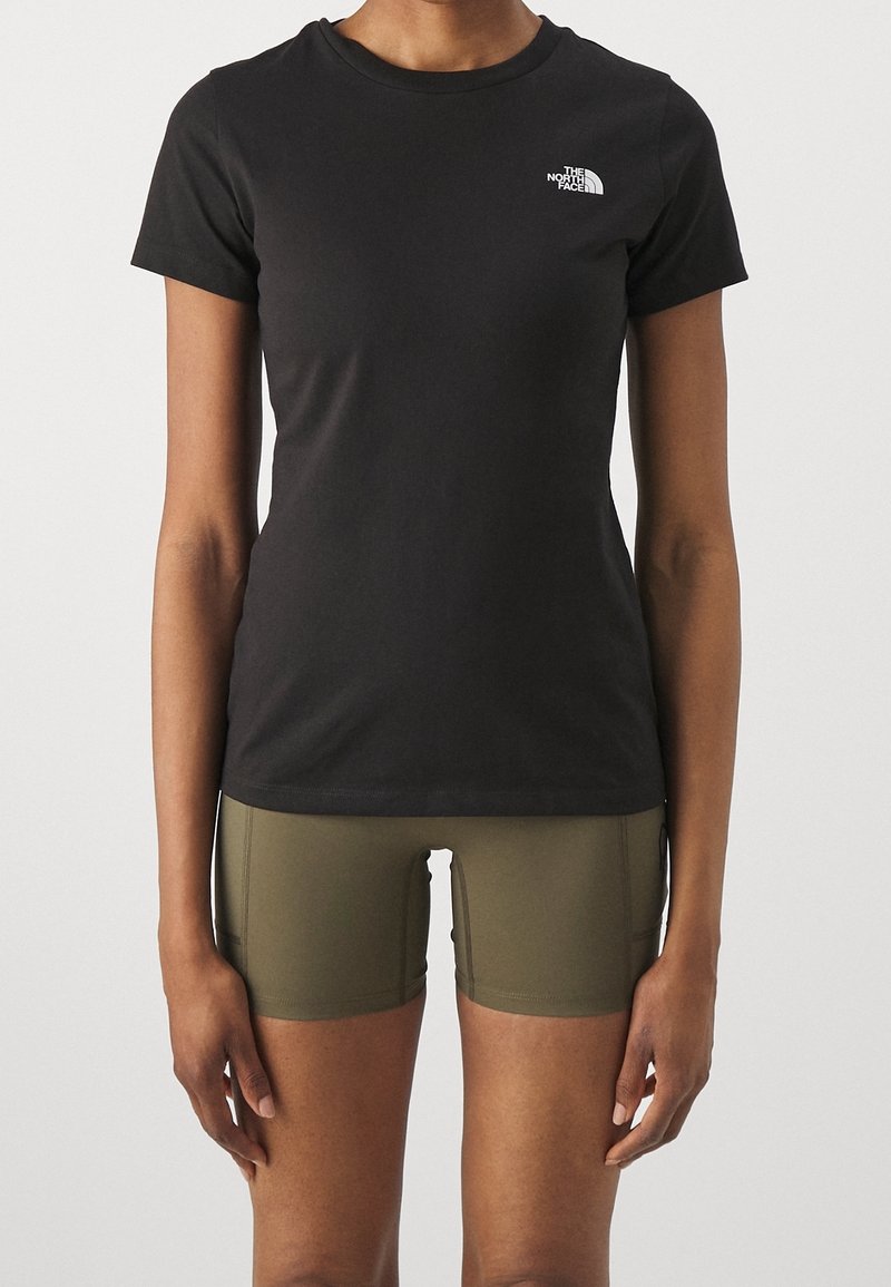 Persona che indossa una t-shirt nera aderente di The North Face e pantaloni corti sportivi verde oliva, in piedi contro uno sfondo chiaro e semplice.