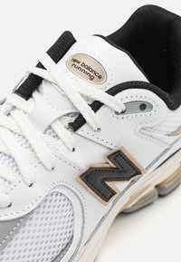 Bílé kožené běžecké boty s mřížkovými panely, černými a šedými akcenty, texturovanými tkaničkami a viditelnou loga nálepkou v béžové barvě.