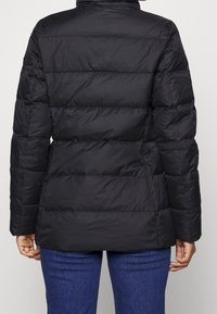 Musta toppatakki, jossa vaakasuora tikkaus, korkea kaulus ja eristetty täyte. Takissa on pitkät hihat ja suora leikkaus, helma ulottuu lantiolle.