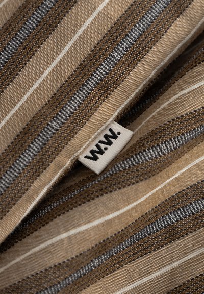 Beżowa tkanina z brązowo-szarymi tkanymi paskami, z małą białą metką z napisem "W.W.", przyszytą na szwie.