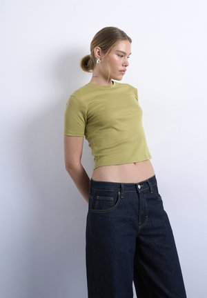 Frau mit hellbraunem Haar, das zu einem niedrigen Dutt gebunden ist, trägt ein hellgrünes, kurzes T-Shirt und dunkelblaue, hoch taillierte Jeans, steht vor einer schlichten weißen Wand.