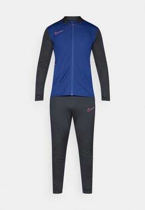 Tuta sportivo da uomo con giacca azzurra con zip e maniche nere, pantaloni coordinati, caratterizzato da loghi Nike rosa sul petto e sulla coscia.