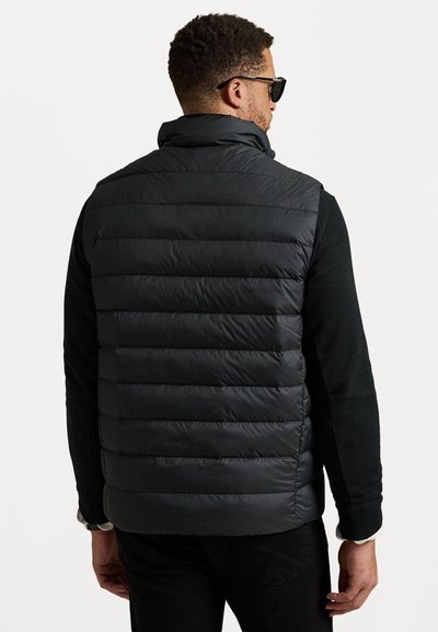 Gilet en duvet noir avec des coutures horizontales, col montant et texture lisse, porté par-dessus une chemise à manches longues noire. Lunettes de soleil incluses.