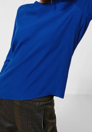Top lungo blu reale a maniche lunghe con vestibilità rilassata e orlo leggermente curvato. Il tessuto sembra morbido con una texture minima. Abbinato a pantaloni a quadri.
