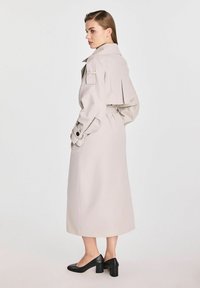 Jasnobeżowy płaszcz trencz z paskiem w talii, dużym kołnierzem i akcentami na ramionach. Noszony na czarnych butach na słupku. Gładka tekstura.